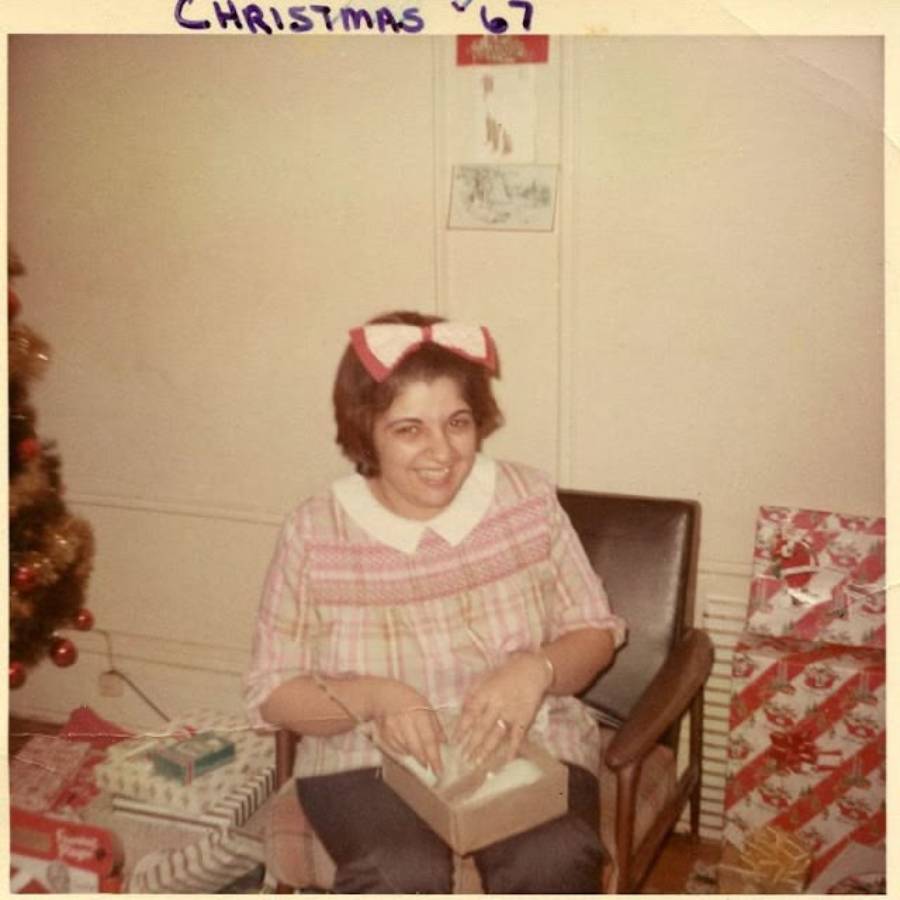 A woman with a pink bow on her head smiles while opening a gift, surrounded by wrapped presents next to a Christmas tree. "Christmas '67" is handwritten at the top of the photo.