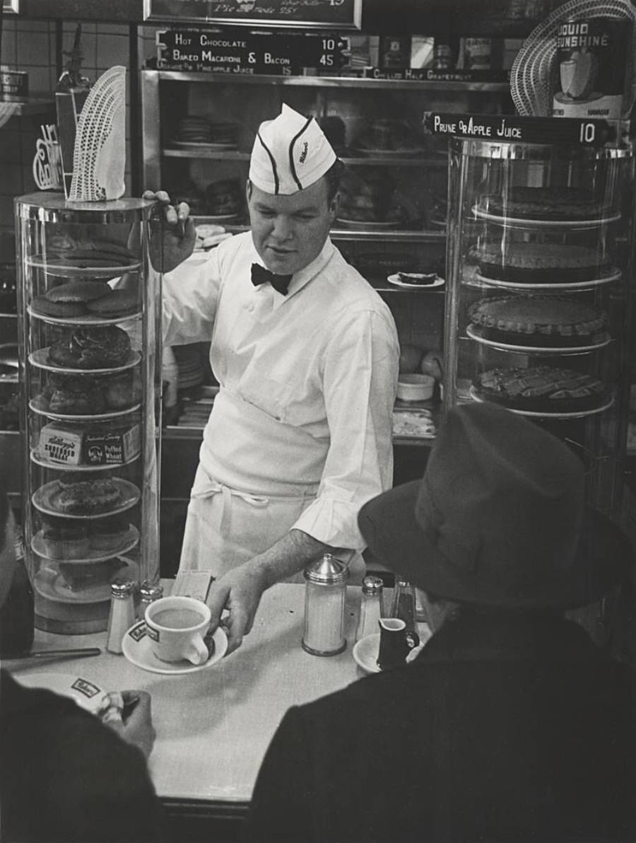 A diner employee in a white uniform serves coffee to two customers seated at a counter, with pastries displayed in glass cases and a menu board in the background.