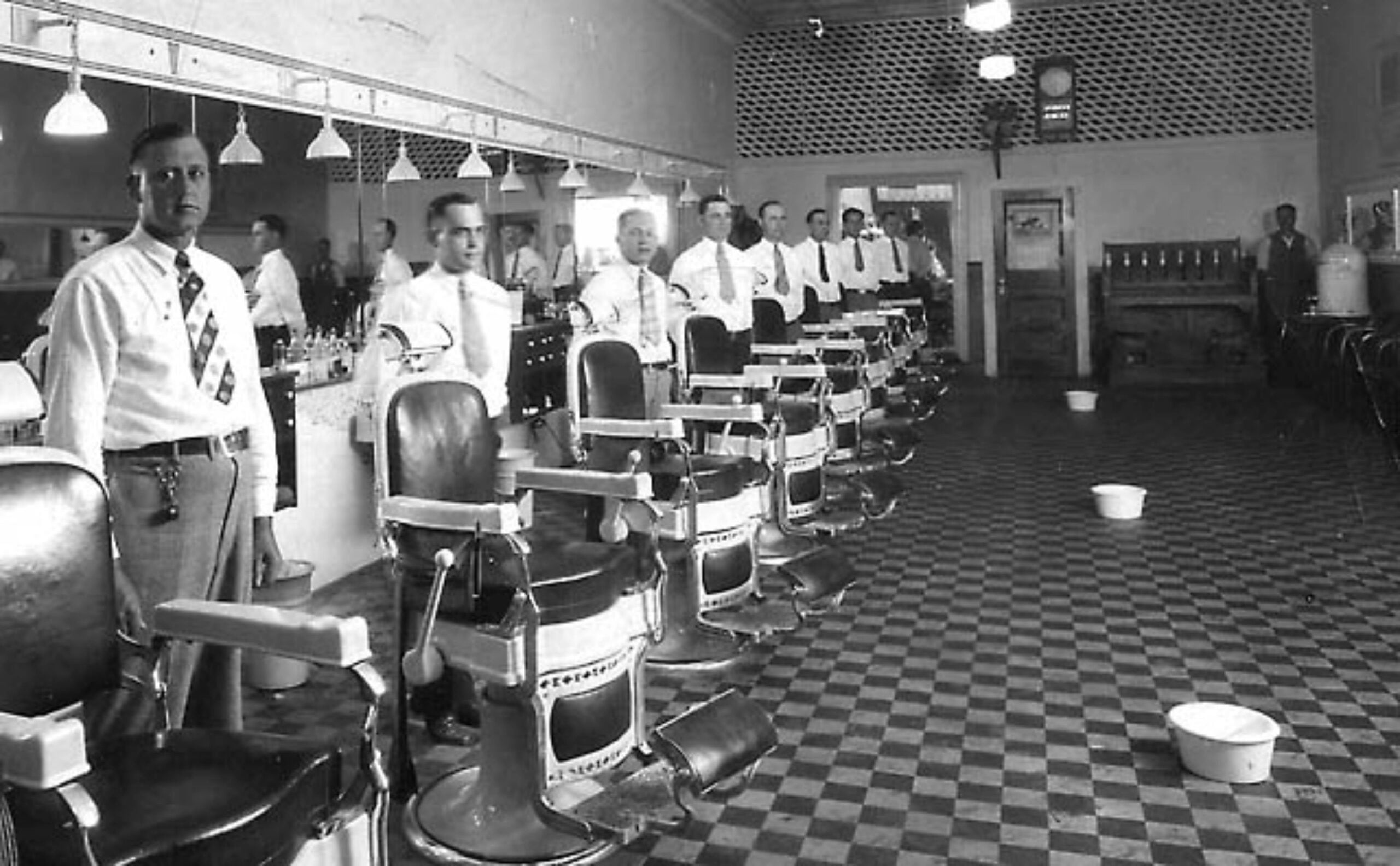 Black and white photo of barbers standing beside empty barber chairs in a vintage barbershop with checkered floor and bowls on the ground.