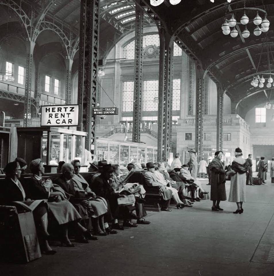 Black and white photo of a busy train station waiting area, with people sitting on benches and a “Hertz Rent a Car” booth visible under a high arched ceiling with large windows.