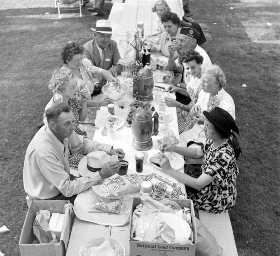 A group of people sit around a picnic table outdoors, eating and talking. The table is covered with food, snacks, and drinks. Some people wear hats and patterned clothing. It appears to be a casual gathering on a sunny day.