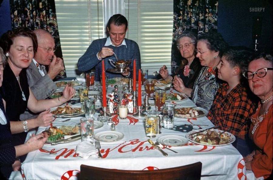 Eight people gather around a table set with food and Christmas decorations, enjoying a holiday meal together. Red candles, festive tablecloth, and plates of food are visible. Some people are smiling, eating, or talking.