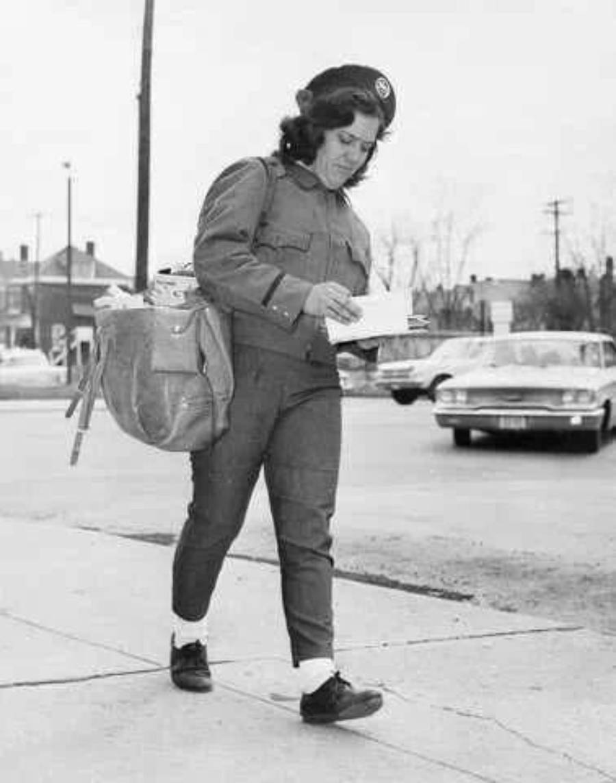 A woman in uniform, carrying a mailbag and reading a letter, walks on a sidewalk in a residential area with cars and houses in the background.