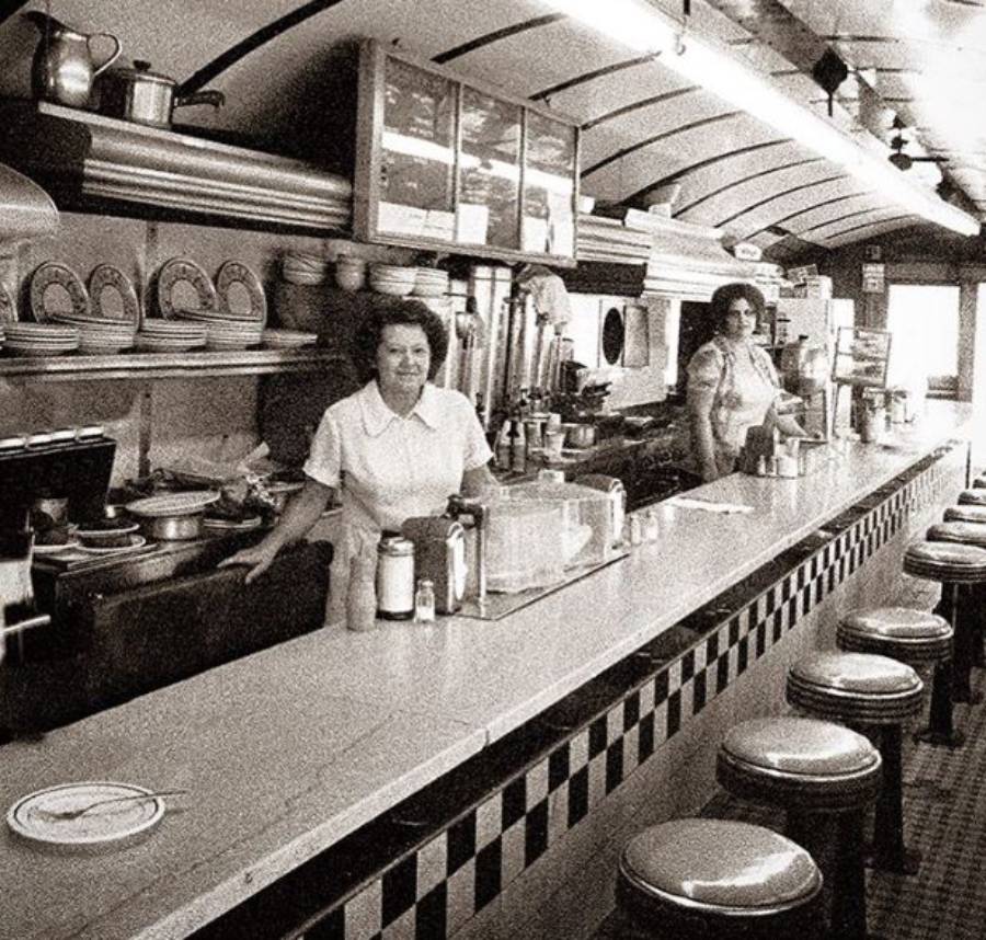 Two women stand behind the counter of a vintage diner with checkered tiles, stools, and shelves of dishes. The diner appears clean and well-lit, with various kitchen items visible in the background.