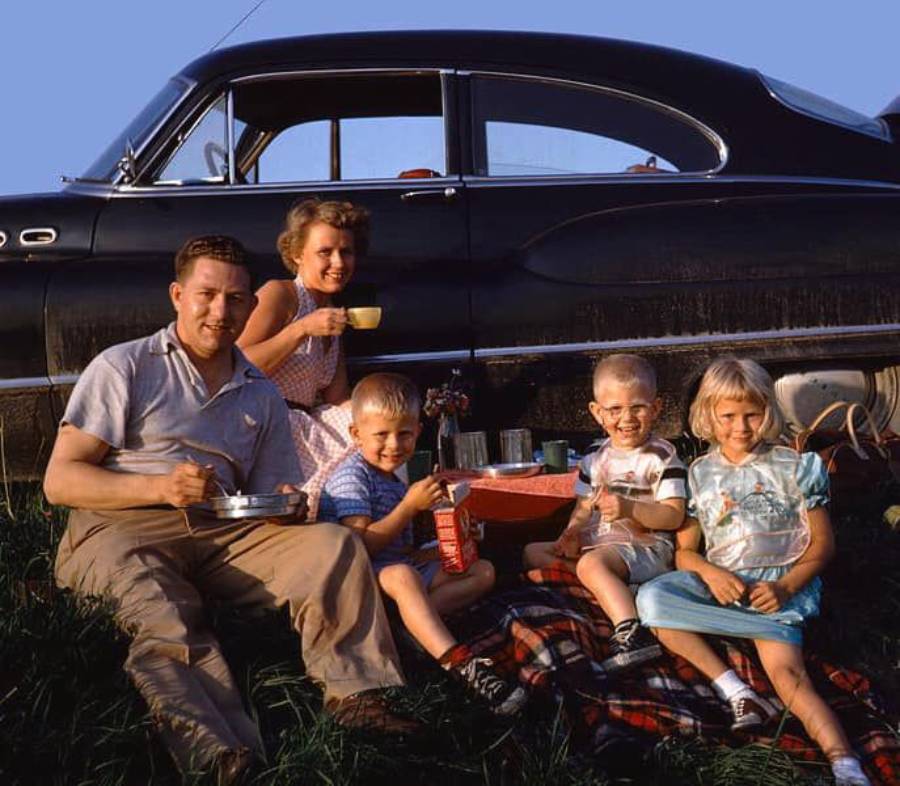 A smiling family of five has a picnic on a blanket next to a vintage black car, enjoying food and drinks outdoors on a sunny day.
