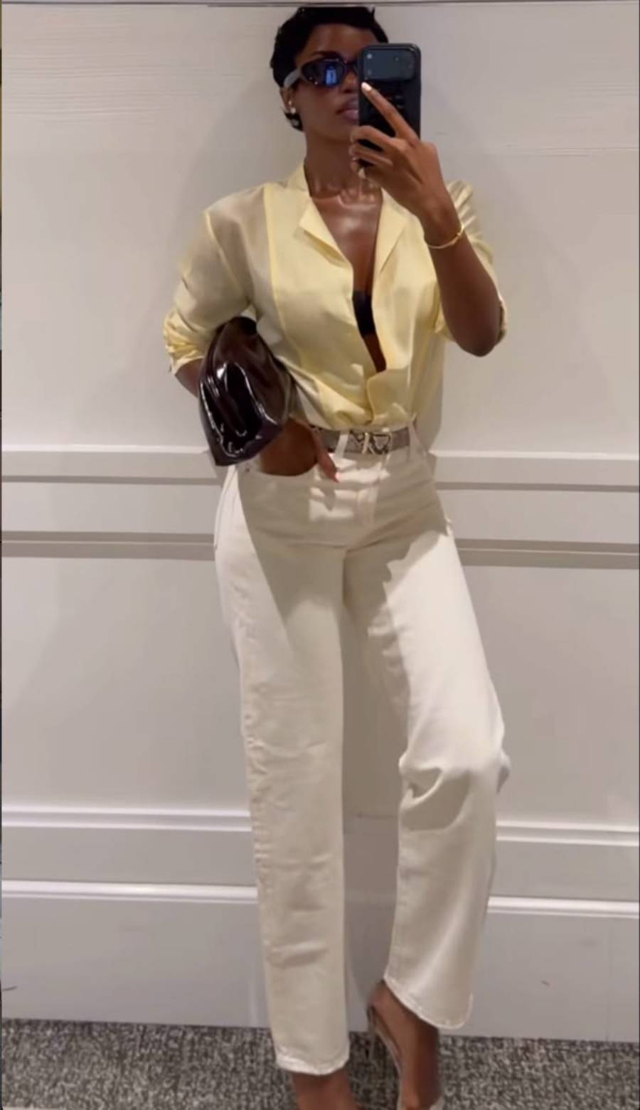 A person wearing sunglasses, a sheer yellow shirt over a black top, white high-waisted pants, and nude heels takes a mirror selfie while holding a dark clutch bag.