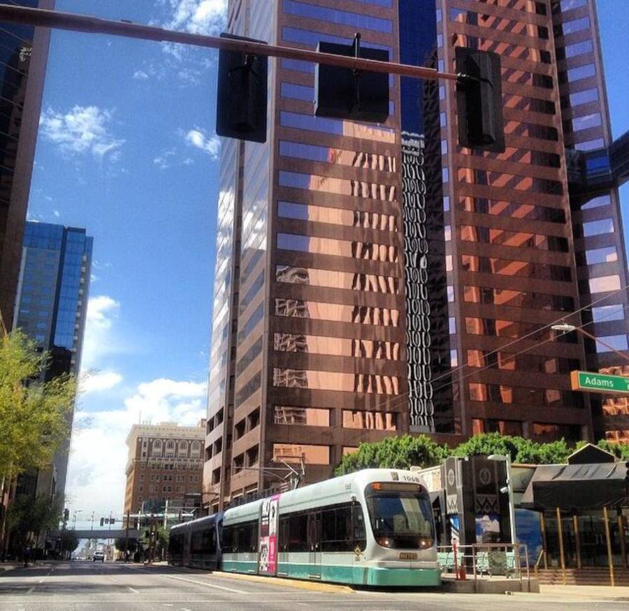 A modern light rail train passes through a city intersection with tall glass buildings under a blue sky. The street signs read "1st Ave" and "Adams St." Traffic lights hang above the scene.