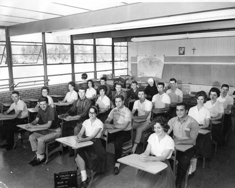 Black and white photo of a classroom with students sitting at desks in rows, facing the camera. A nun is seated among them. Large windows line one wall, and a map and crucifix are visible on the back wall.