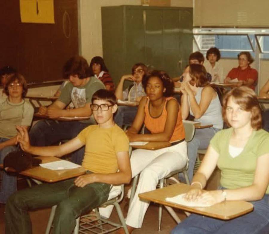 A classroom of high school students sit at desks, some looking at the camera, others focused elsewhere. The scene looks like it’s from the 1970s, with retro clothing and hairstyles.