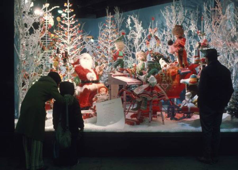 People admire a festive holiday display with Santa Claus, elves, and toys among decorated Christmas trees and snow. Two adults and a child are silhouetted in front of the brightly lit, colorful scene.