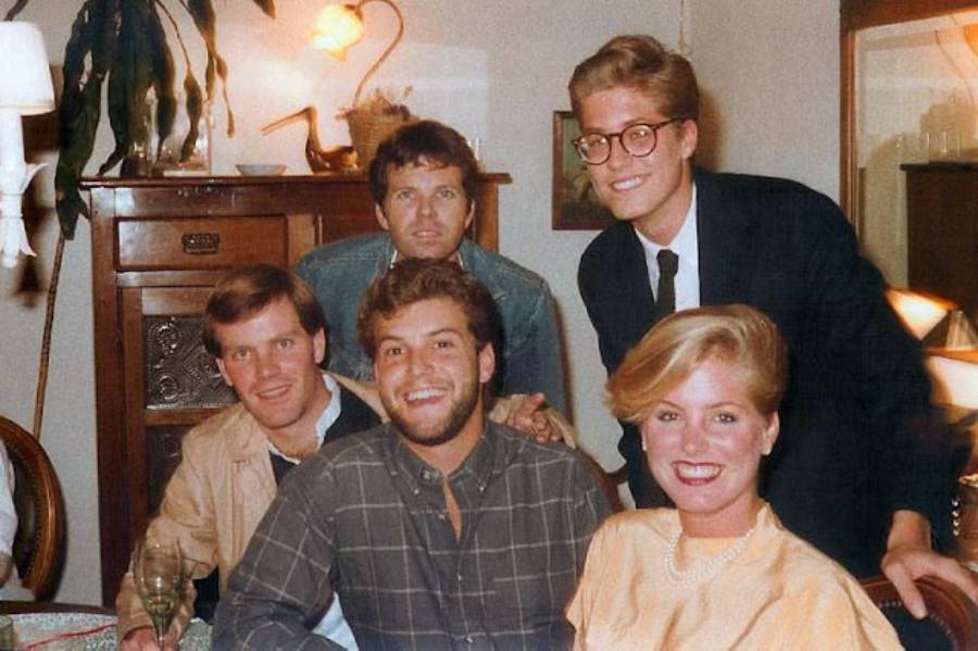 Five young adults, four men and one woman, pose and smile indoors around a table. There is a wooden cabinet and lamp in the background, and the group appears happy and casually dressed.