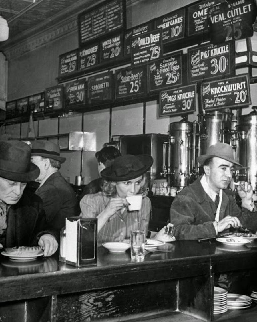 Black and white photo of people sitting at a diner counter, eating and drinking. Behind them are coffee urns and a wall menu listing food prices. All are wearing hats and jackets, suggesting a past era.