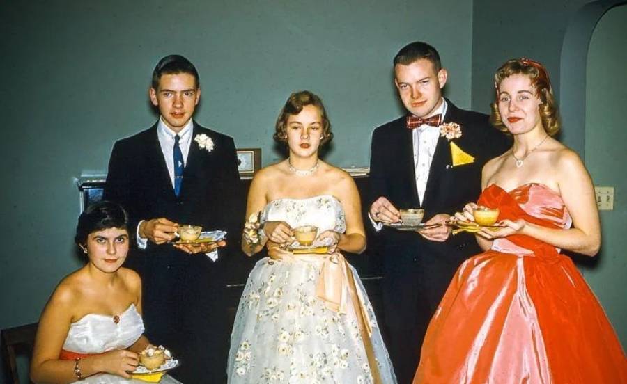 Five young adults dressed in formal 1950s attire pose indoors, holding plates with desserts. Three women wear elegant strapless gowns, while two men wear suits with bow ties and boutonnieres.