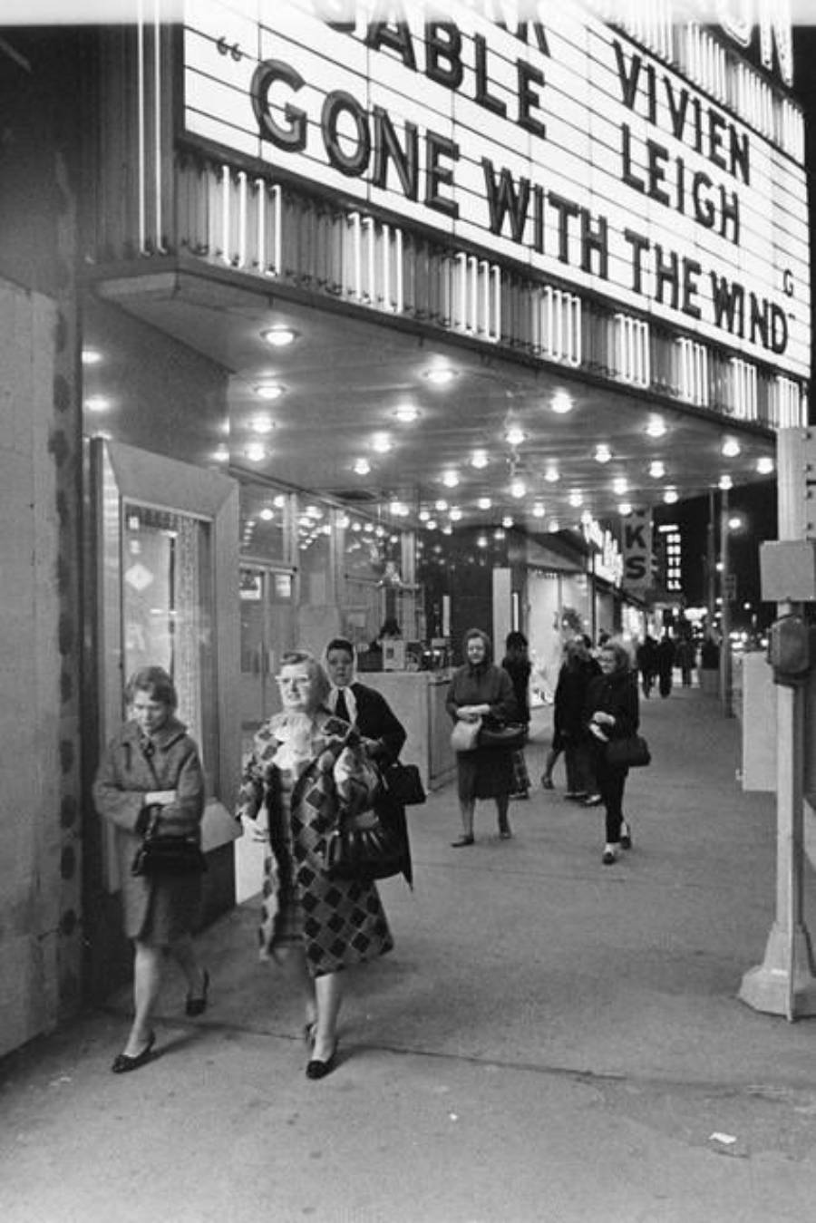 Black-and-white photo of people walking outside a movie theater at night. The marquee advertises "Gone With the Wind" starring Clark Gable and Vivien Leigh. Bright lights illuminate the sidewalk and theater entrance.