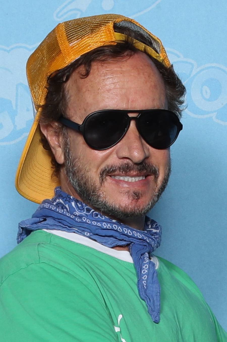 A man wearing a yellow cap backwards, black sunglasses, a blue bandana around his neck, and a green shirt, smiling in front of a light blue background with white designs.