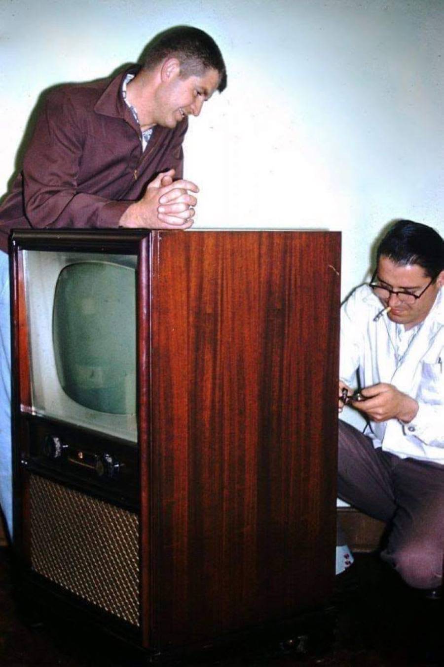 Two men are next to a large vintage wooden television; one stands leaning on it while the other sits beside it, adjusting or examining something in his hands.