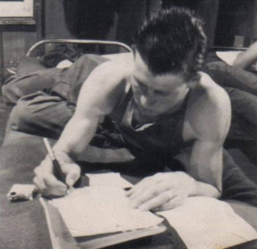 A man in a sleeveless shirt lies on a bed, leaning on his elbow while writing with a pencil on a sheet of paper. Papers and a small item are on the bed beside him. The setting appears to be a simple, indoor room.