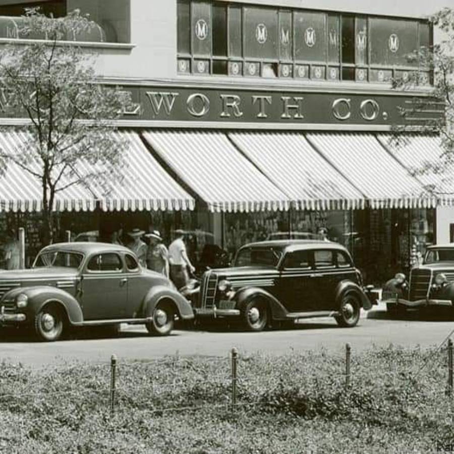 Black and white photo of vintage cars parked outside an F.W. Woolworth Co. store with striped awnings, people walking on the sidewalk, and large display windows above the entrance.