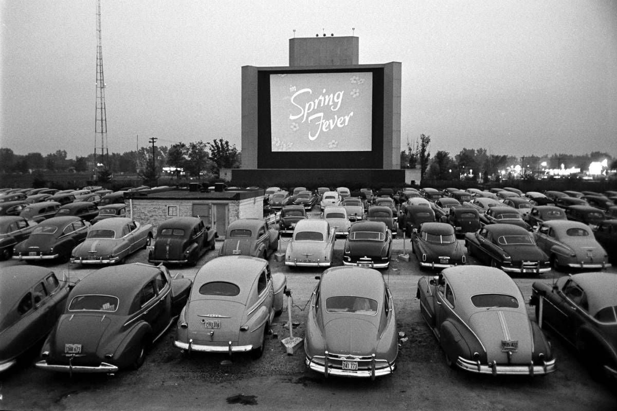 Black-and-white photo of a drive-in theater filled with vintage cars facing a large screen that displays the title "Spring Fever" at dusk, with trees and lights visible in the background.