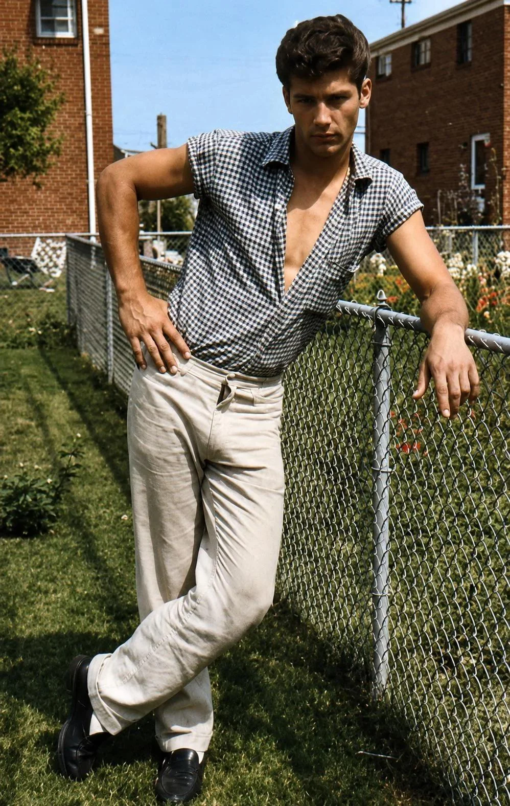 A man in a checkered short-sleeve shirt and light pants leans against a chain-link fence in a grassy yard, with brick buildings in the background.