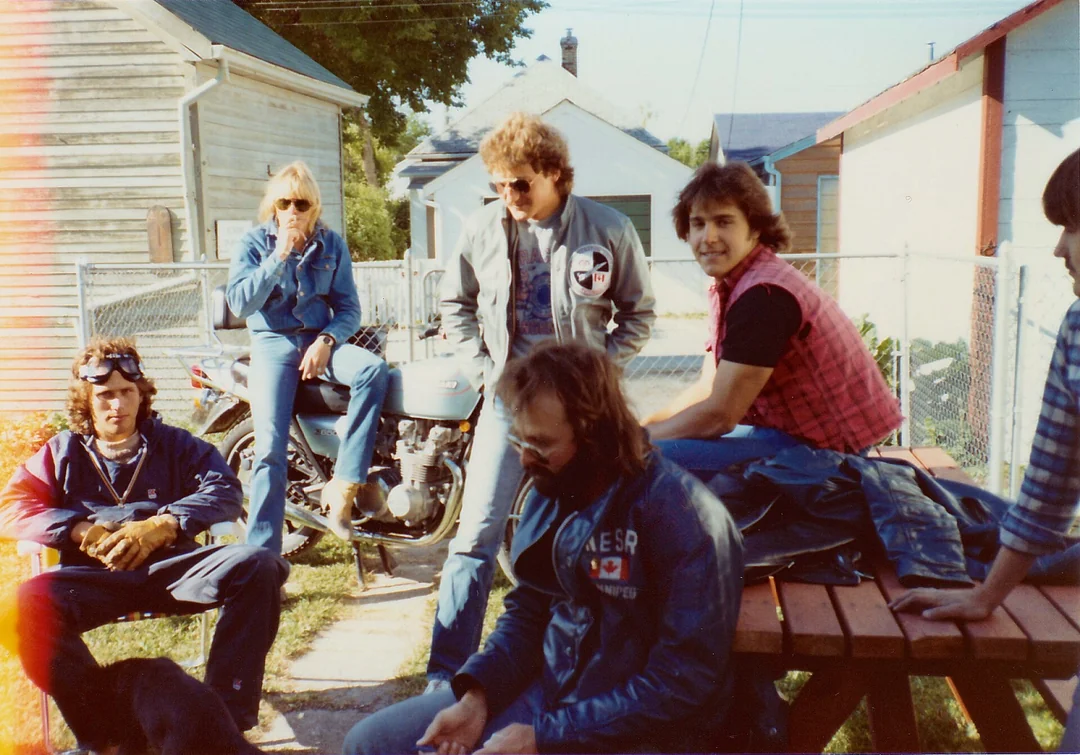 Five people in casual 1980s attire relax outdoors; two sit on a picnic table, two stand, and one sits on the grass near a motorcycle, with a house and fence in the background.
