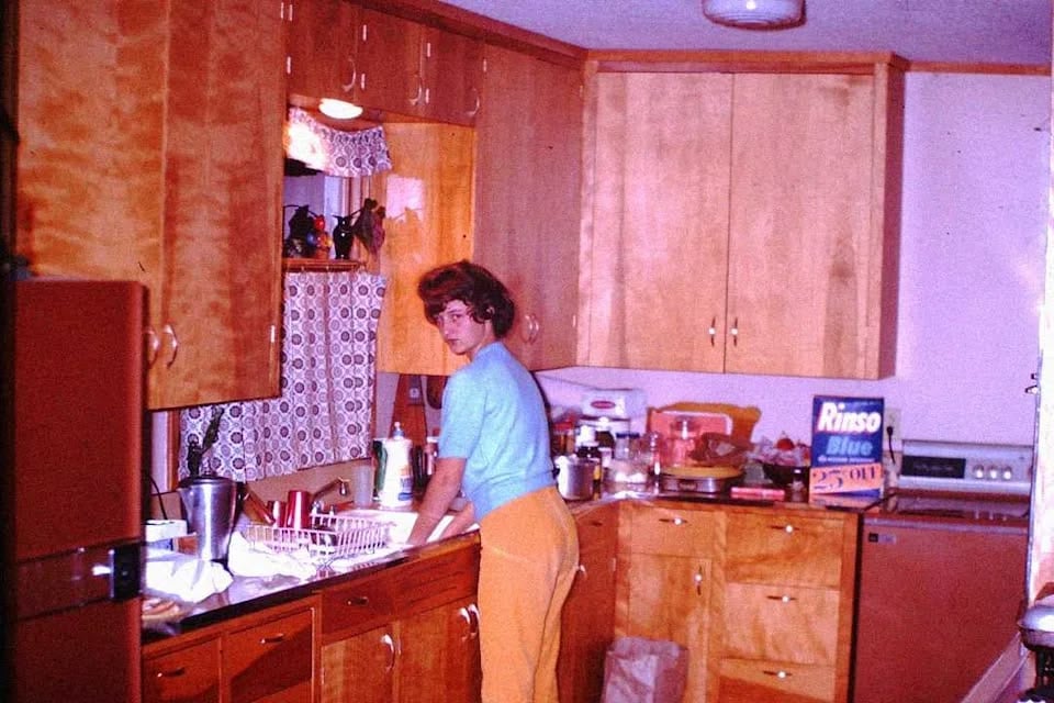 A woman stands at a kitchen sink washing dishes. The kitchen has wooden cabinets, a dish rack, and a Risso Blue detergent box on the counter. The scene appears to be from the mid-20th century.