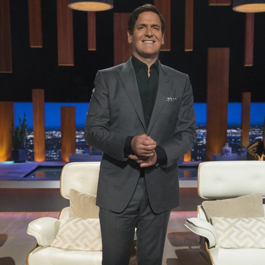 A man in a gray suit stands smiling in a modern studio set with white chairs, pillows, and cityscape visible through large windows in the background.