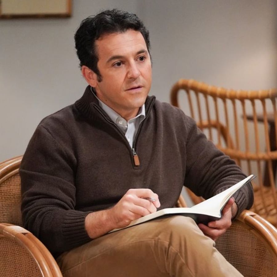 A man sits in a wicker chair, holding a pen and open notebook, wearing a brown zip-up sweater over a collared shirt and khaki pants, looking attentive, with more wicker chairs in the background.