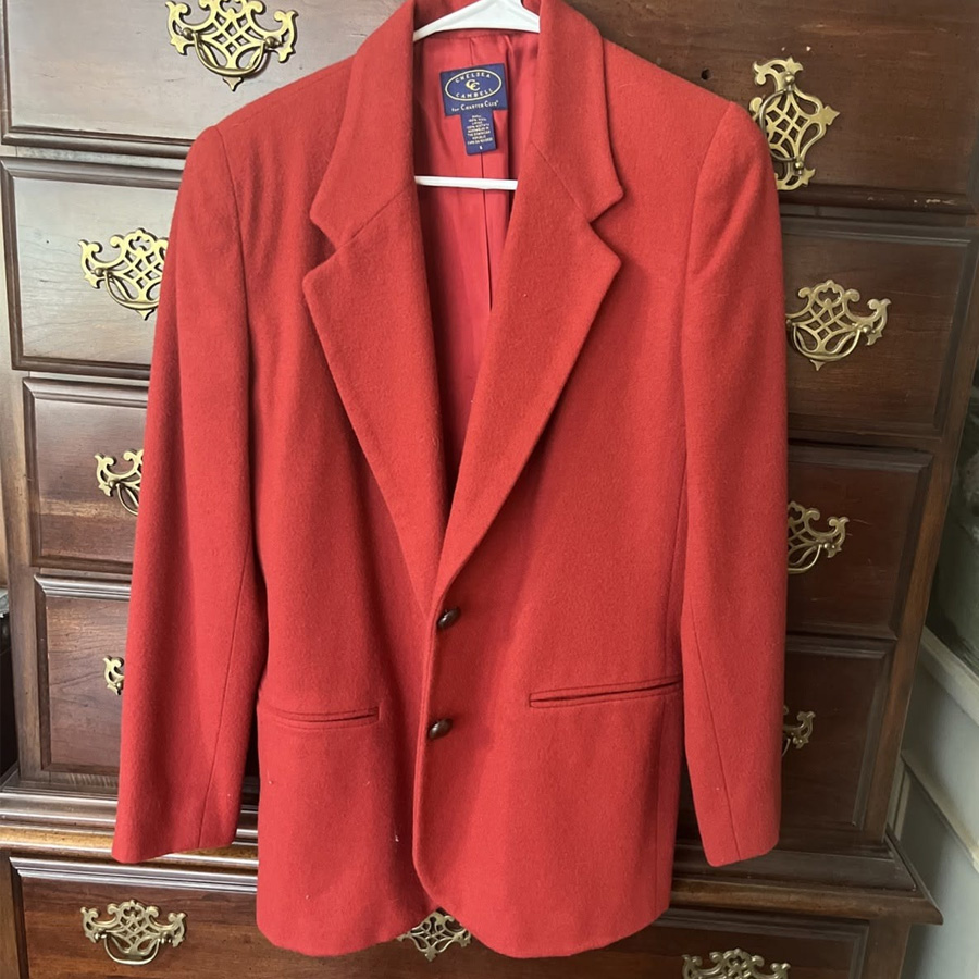 A red wool blazer with two buttons and two front pockets hangs on a wooden dresser with ornate brass handles.