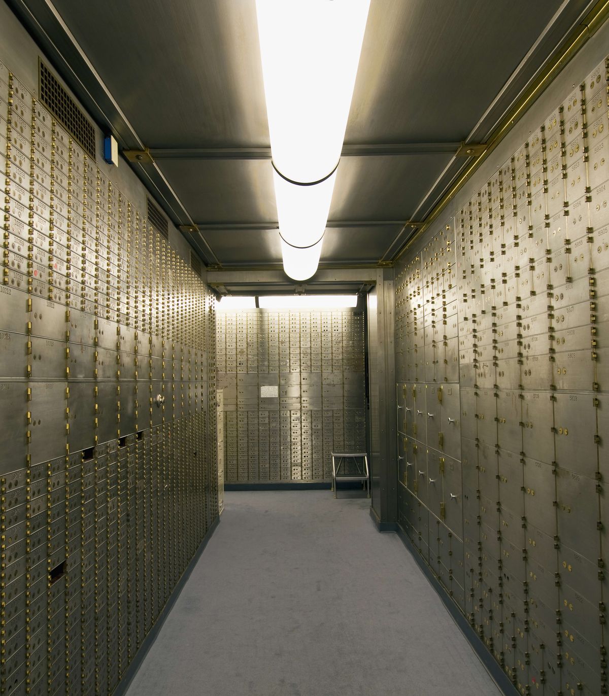 A brightly lit bank vault room with metal walls lined with rows of gold-colored safety deposit boxes and a small table at the end of the room.
