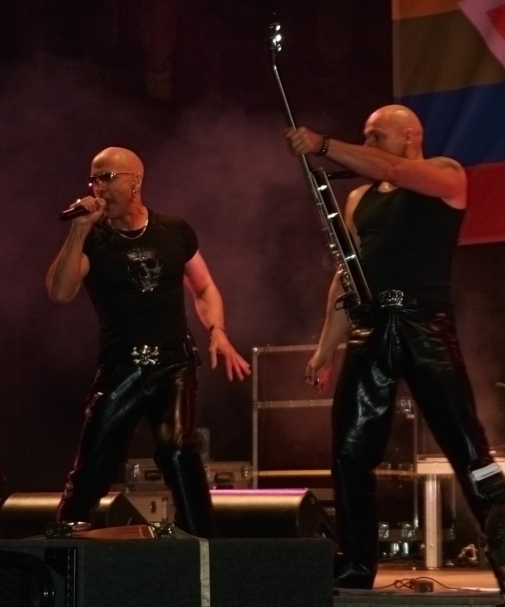 Two men in black shirts and shiny black pants perform on stage, one singing into a microphone and the other playing a guitar. They both wear sunglasses and appear energetic under stage lights.