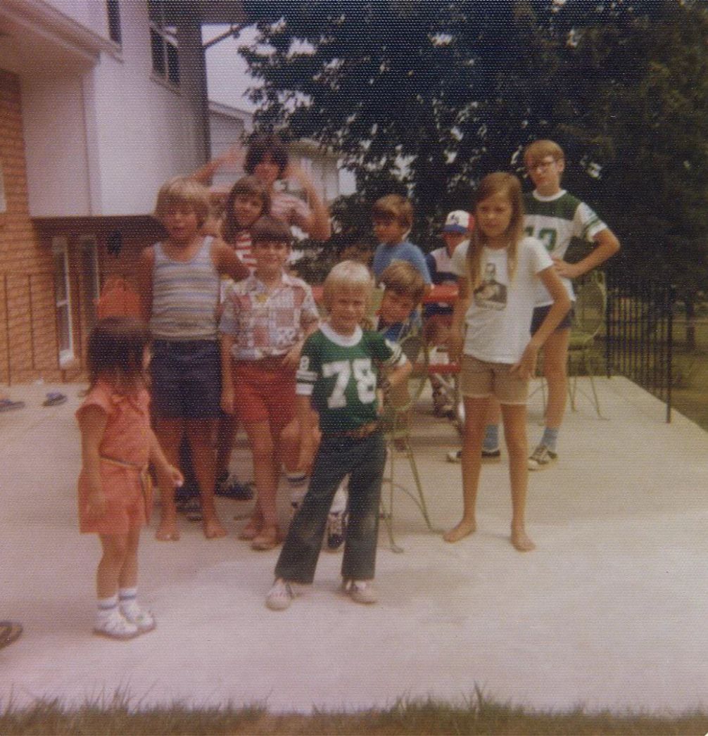 A group of children stand and pose on a concrete patio outside, with some smiling at the camera. One boy in a green "78" jersey stands front and center. Trees and a house are visible in the background.