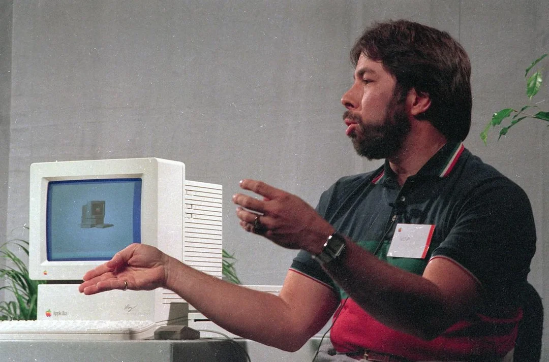 A man gestures while sitting next to a vintage Apple Macintosh computer and disk drives on a table, with a plain background and some green plants nearby.