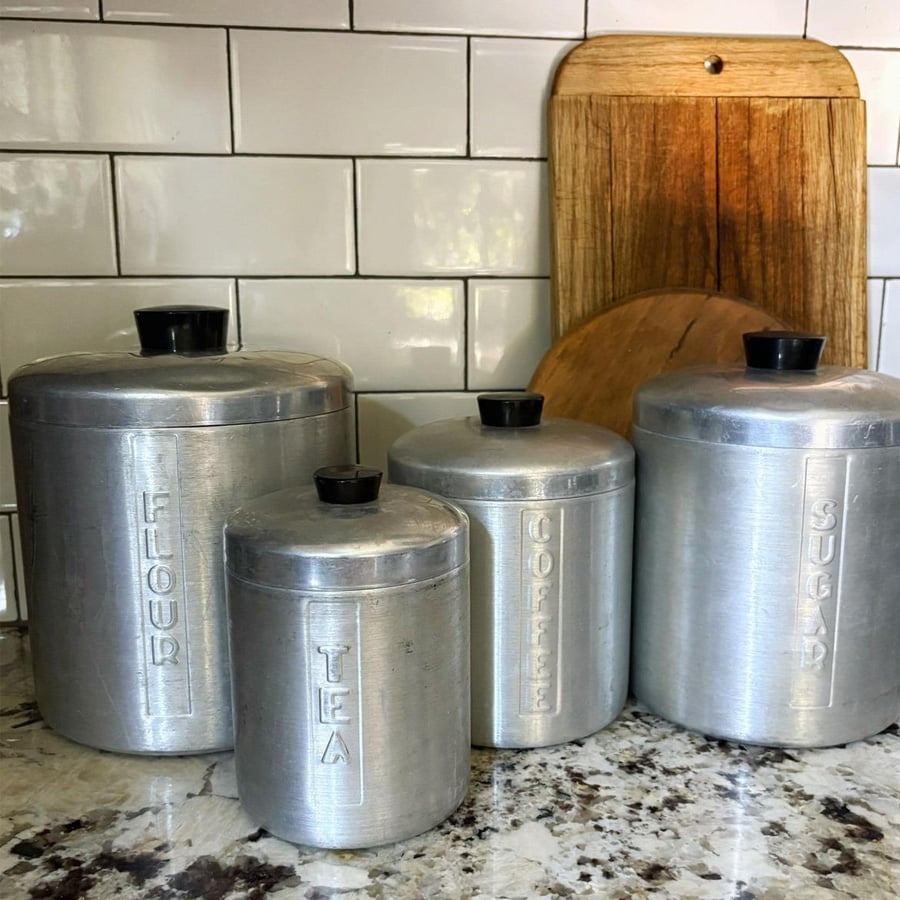 Four metal canisters labeled flour, tea, coffee, and sugar sit on a kitchen counter in front of white subway tiles and two wooden cutting boards.