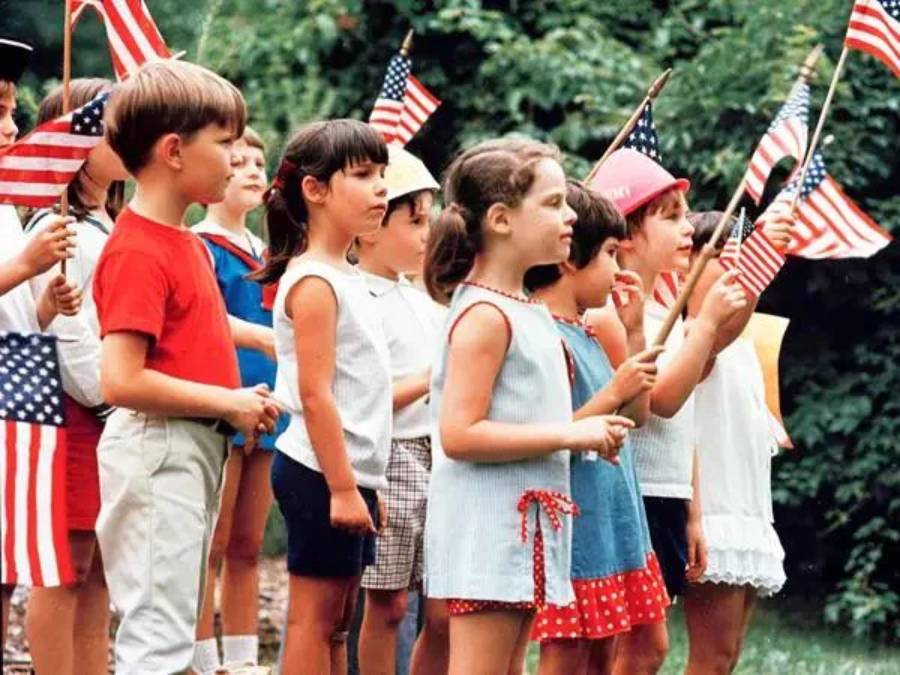 A group of children stand outdoors holding small American flags, dressed in summer clothes, appearing to participate in a patriotic event or celebration. Green trees are visible in the background.