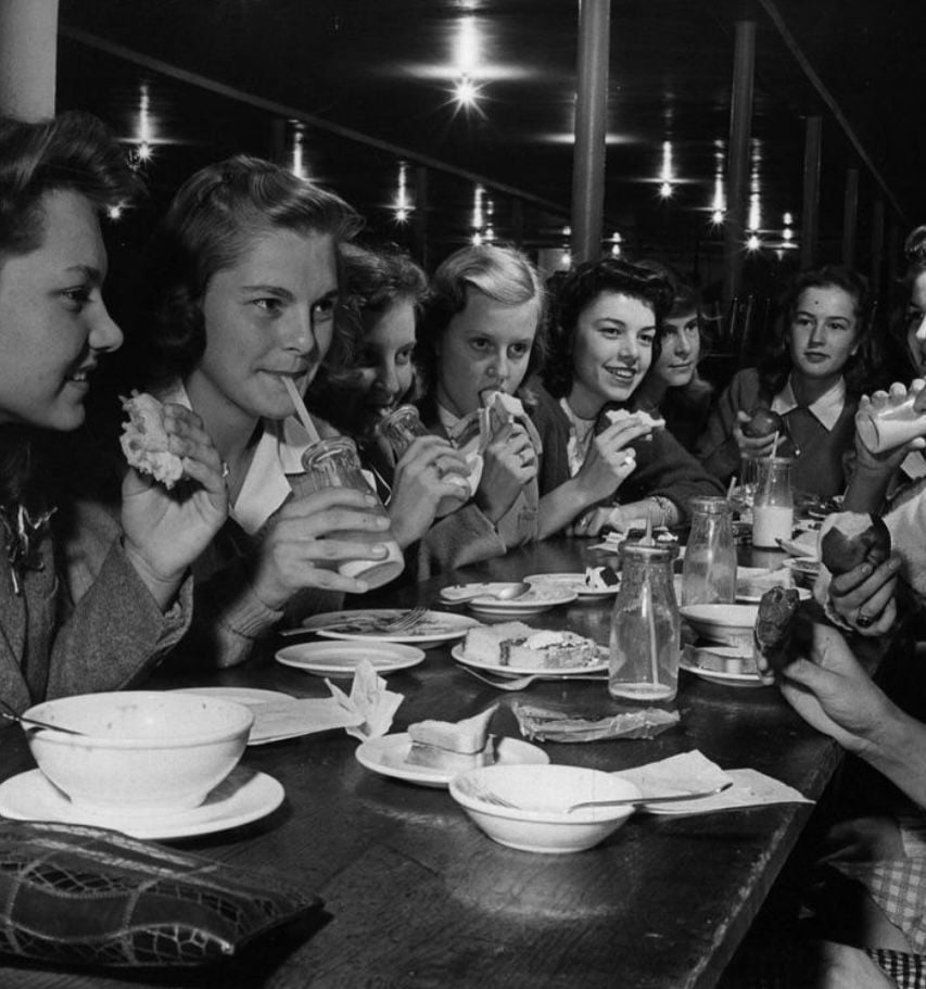 A group of young women sit closely together at a long table, smiling and eating sandwiches and drinks, with plates, bowls, and milk bottles in front of them in a lively indoor setting.