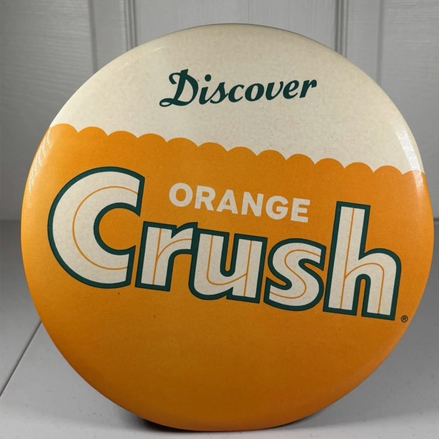 Round button with "Discover Orange Crush" text in green and orange on a two-tone white and orange background, resembling the branding of the Orange Crush soda.