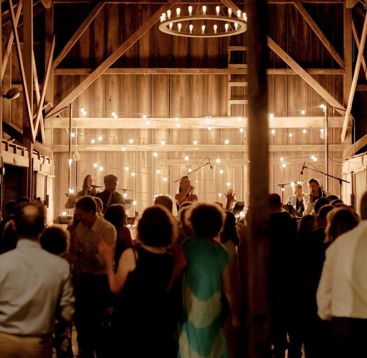 A lively crowd dances in a warmly lit barn, with string lights and a band playing on stage in the background. The atmosphere is festive and inviting.