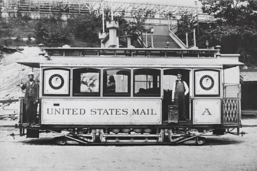 Old black and white photo of a United States Mail streetcar with two men standing at each end, dressed in work clothes, on a dirt road with a stairway and trees in the background.