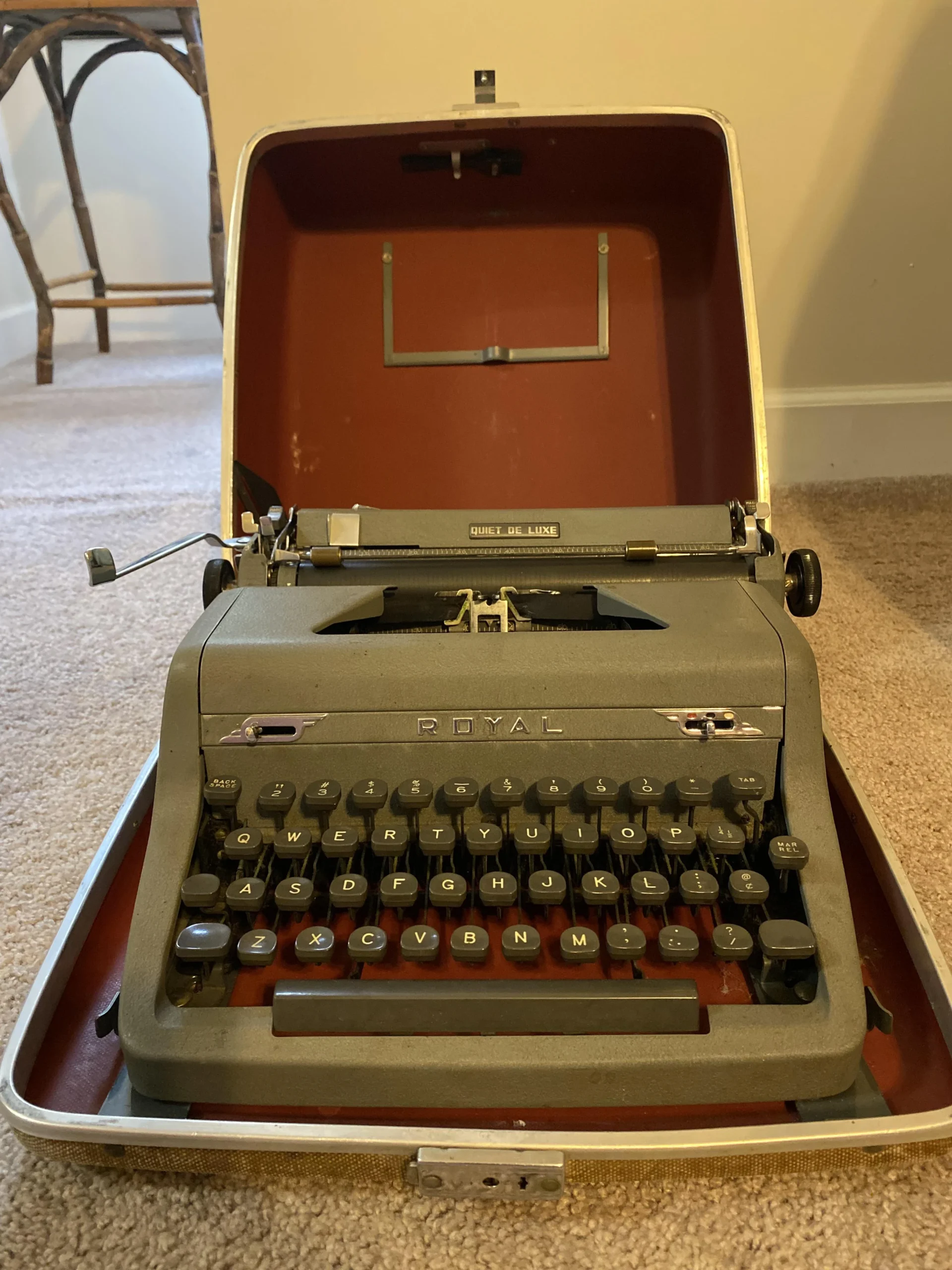A vintage gray Royal typewriter sits in an open hard-shell carrying case on a beige carpet, with a wicker chair and cream-colored wall visible in the background.