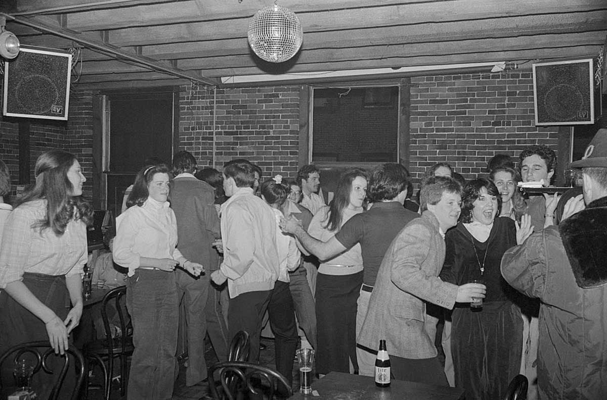 A group of people socialize and dance in a crowded bar with brick walls, wooden ceiling beams, and a disco ball overhead. Some hold drinks and talk, while others stand or move around the room.