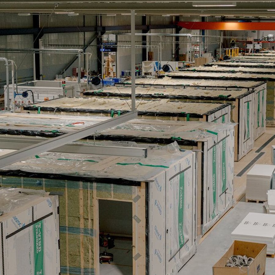 Interior of a factory with partially assembled modular home units lined up in rows, surrounded by construction materials and equipment under bright lighting.