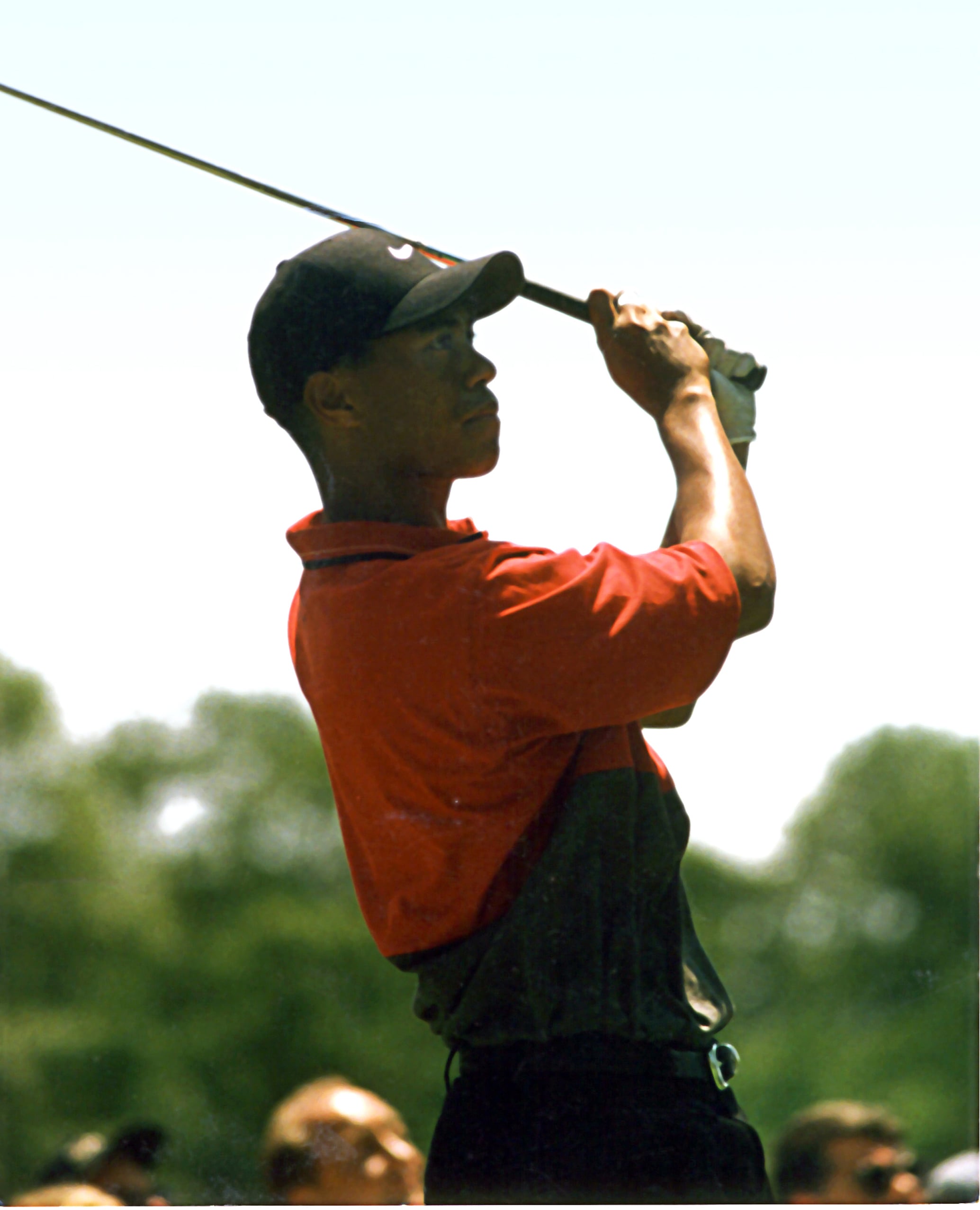 A golfer wearing a red shirt and black cap follows through after hitting a golf ball, with trees and people blurred in the background.