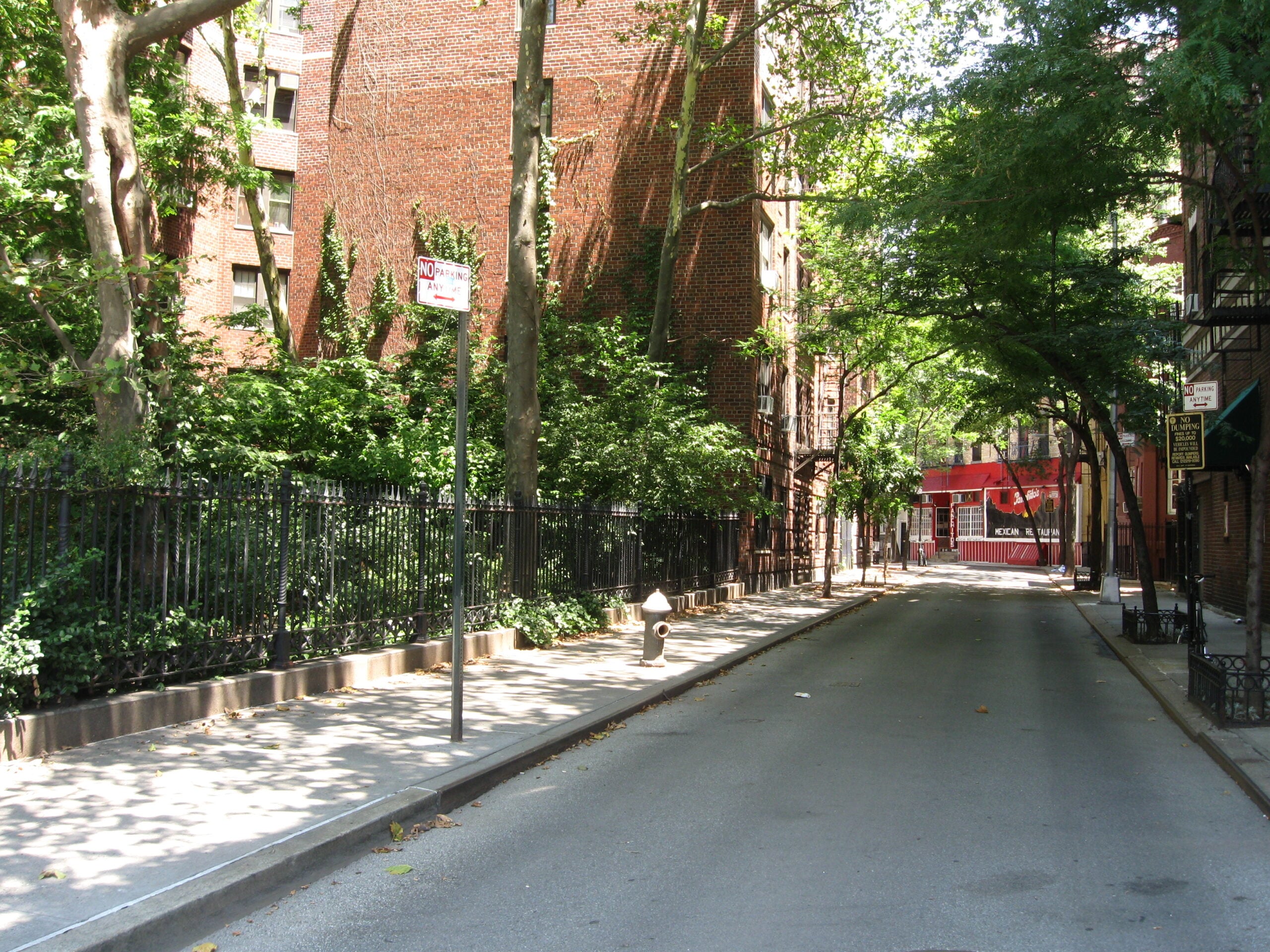 A quiet, empty street lined with trees and brick buildings on a sunny day. Shadows from the trees cover the street, and a red storefront is visible in the background.