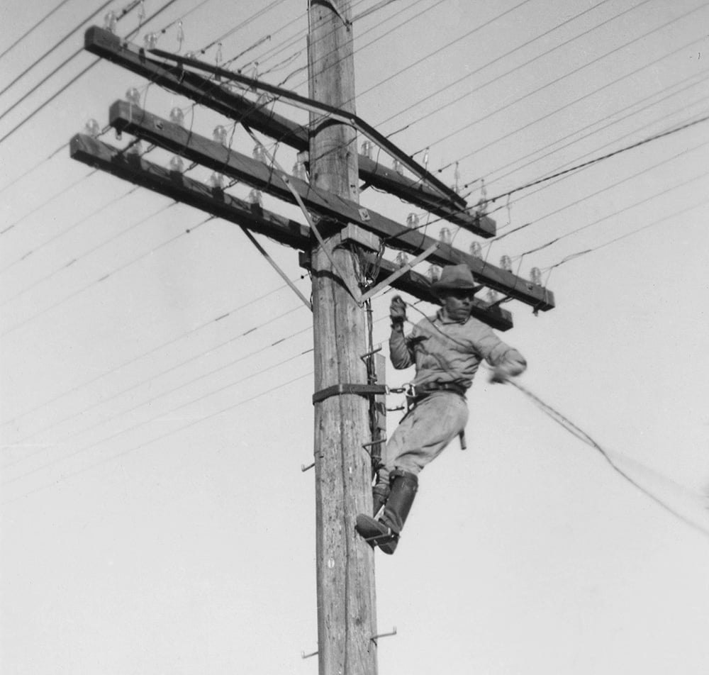 A utility worker in work clothes and boots is suspended from a utility pole, holding onto a rope while performing maintenance on electrical wires high above the ground.