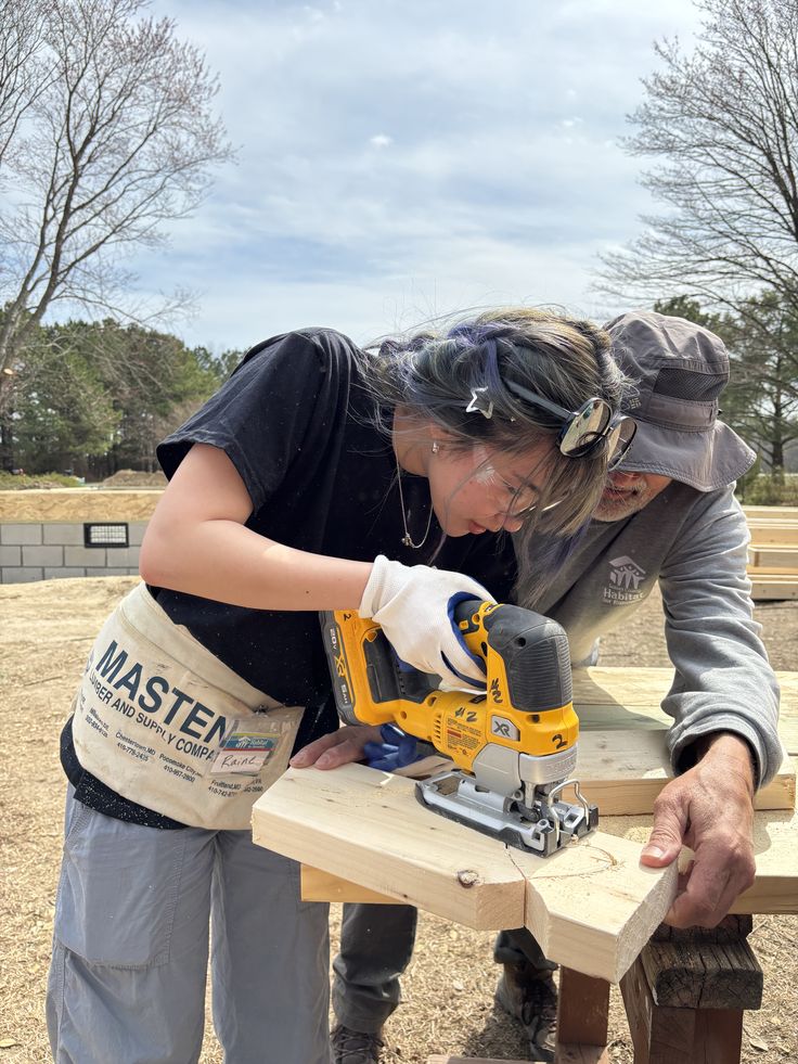 A young woman uses a jigsaw to cut wood while an older man assists her outdoors. Both wear safety glasses, and she has a tool belt on. Trees and building materials are visible in the background.