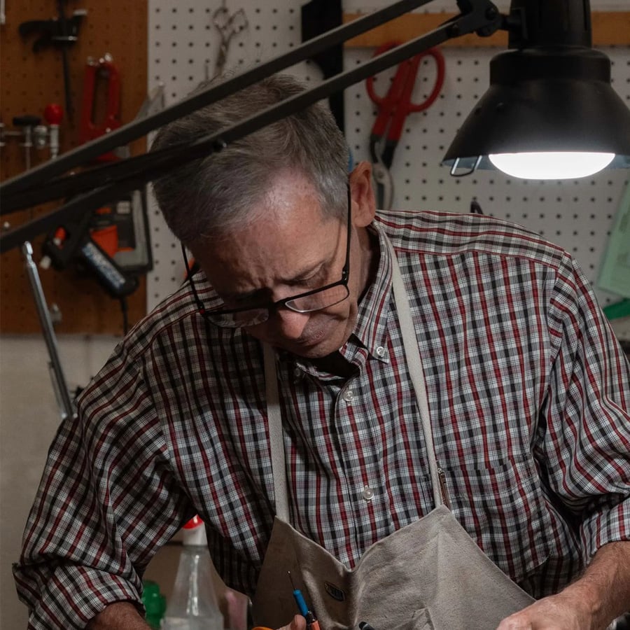 An older man wearing glasses and a plaid shirt works at a workbench under a lamp, surrounded by tools hanging on a wall in a workshop.