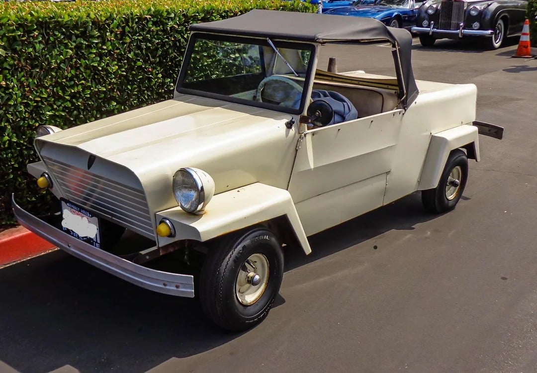 A small, vintage, cream-colored convertible car with a black soft top is parked on a street near a hedge and a red curb. The car has round headlights and a simple, boxy design.