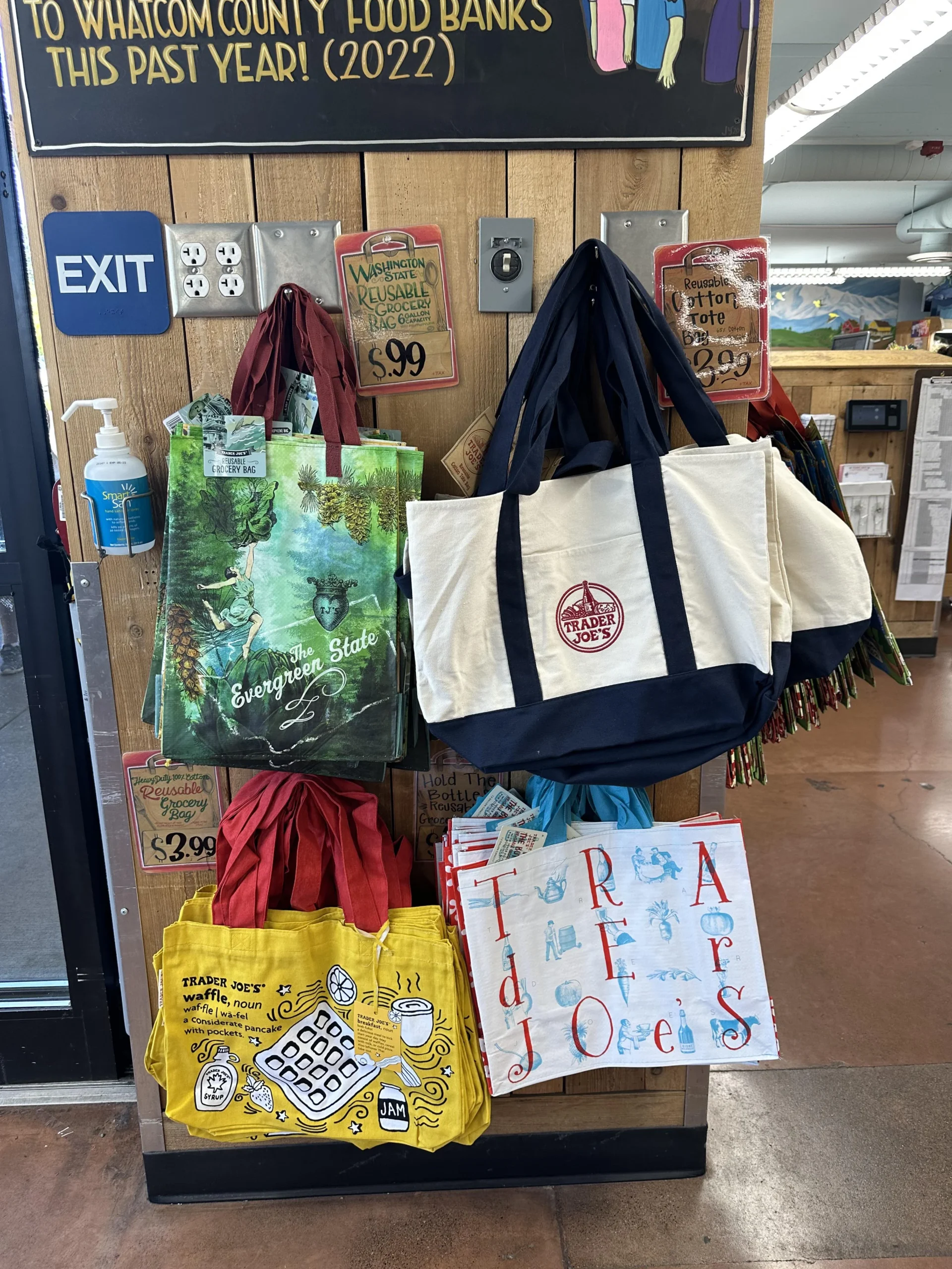 Reusable tote bags hang on display near an exit in a store; designs include an evergreen forest, a cream and navy logo, a crossword puzzle, and colorful hand-drawn letters spelling "Trader Joe’s.