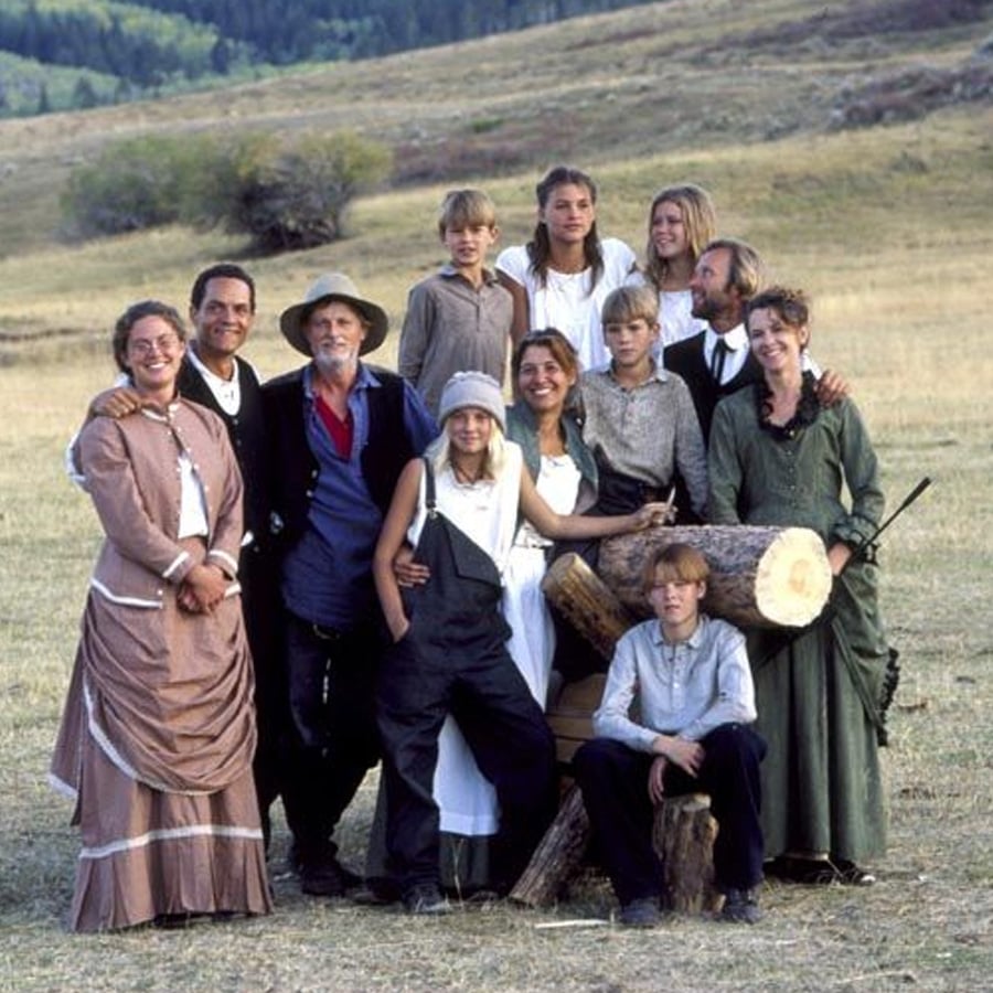 A group of people in pioneer-era clothing pose together outdoors in a grassy field, with logs stacked in front and hills in the background.